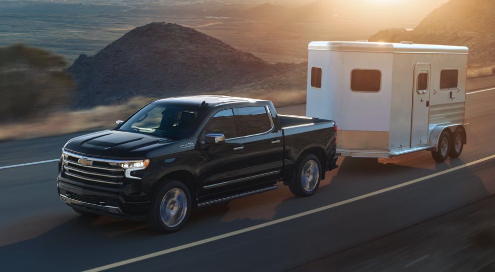 Black 2026 Chevy Silverado 1500 High Country towing a trailer on a paved road.
