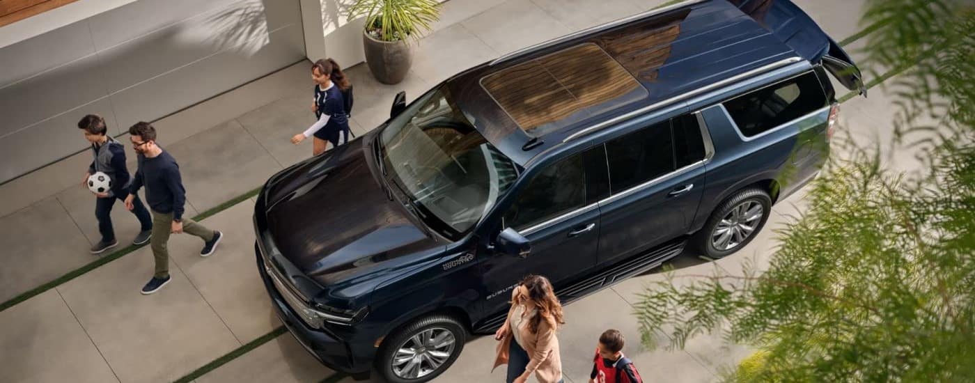 A dark blue 2023 Chevy Suburban High Country is shown from a highway on a driveway near a family.